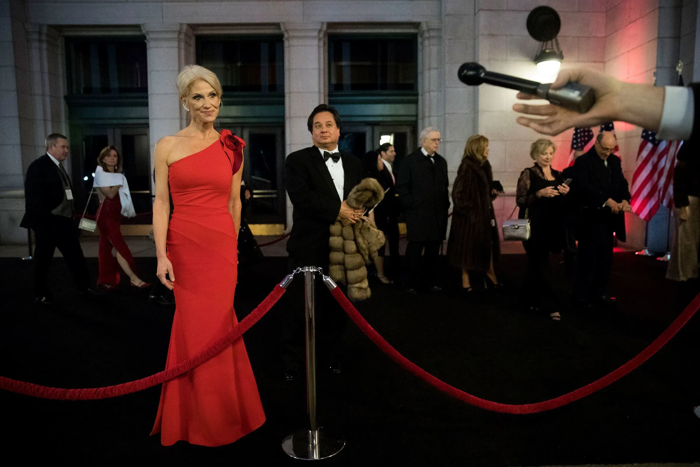 امرأة ترتدي فستانًا أحمر أنيقًا تقف أمام حواجز حمراء في حدث رسمي، بينما يظهر رجال ونساء آخرون في الخلفية.