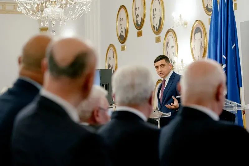 سيميون يتحدث في مؤتمر، مع صور تاريخية معلقة على الجدران، وسط مجموعة من الحضور الذين يستمعون له.