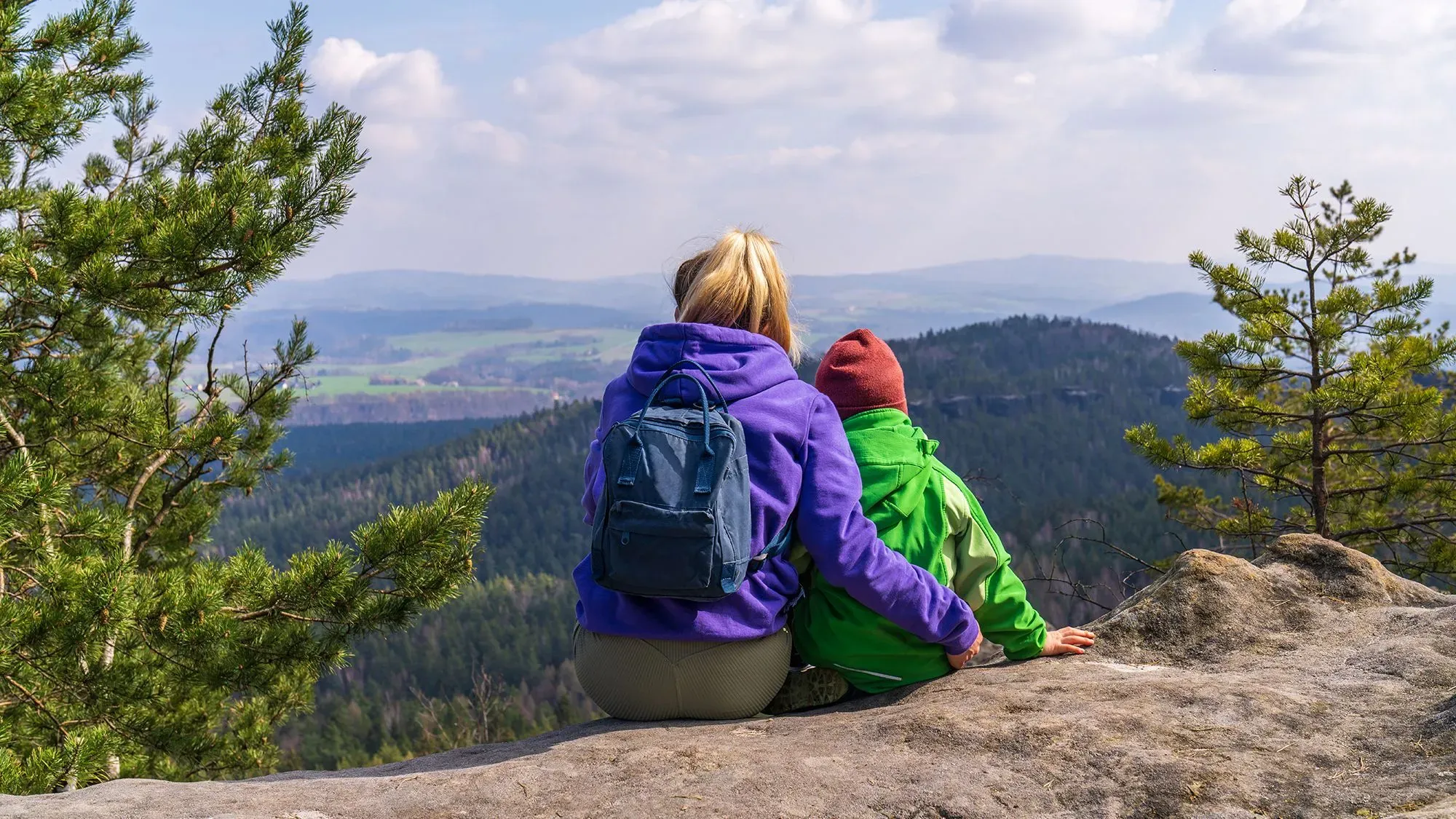 امرأة وطفل يجلسان معًا على حافة صخرية، يستمتعان بالمنظر الطبيعي الخلاب للجبال والغابات، مما يعكس أهمية الحرية والرفاهية.