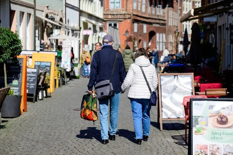 زوجان يسيران معًا في شارع مزدحم، يحملان أكياس تسوق، مع مقاهي ومحلات تجارية في الخلفية، مما يعكس الحياة اليومية في المدينة.