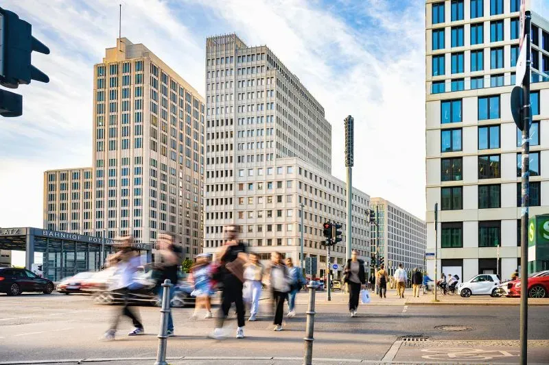 مشهد حضري يظهر أشخاصًا يعبرون الشارع أمام مباني حديثة في برلين، مما يعكس الحياة النشطة في المدينة وتأثير الدخل الأساسي على المجتمع.