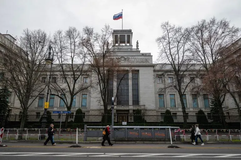 مبنى السفارة الروسية في برلين مع العلم الروسي يرفرف فوقه، بينما يسير بعض الأشخاص على الرصيف في أجواء مشحونة بالتوتر الدبلوماسي.