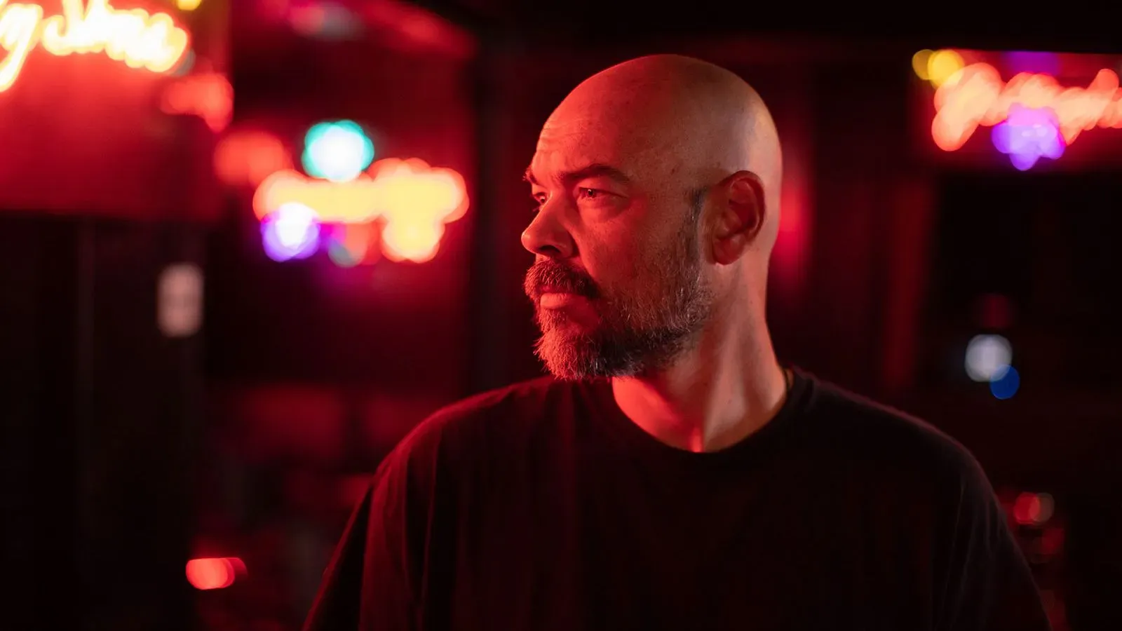 تظهر الصورة رجلًا ذو لحية وشعر أصلع، يقف في مكان مضاء بأضواء نيون ملونة، مع تعبير جاد يعكس التوتر المرتبط بقضية جريمة.