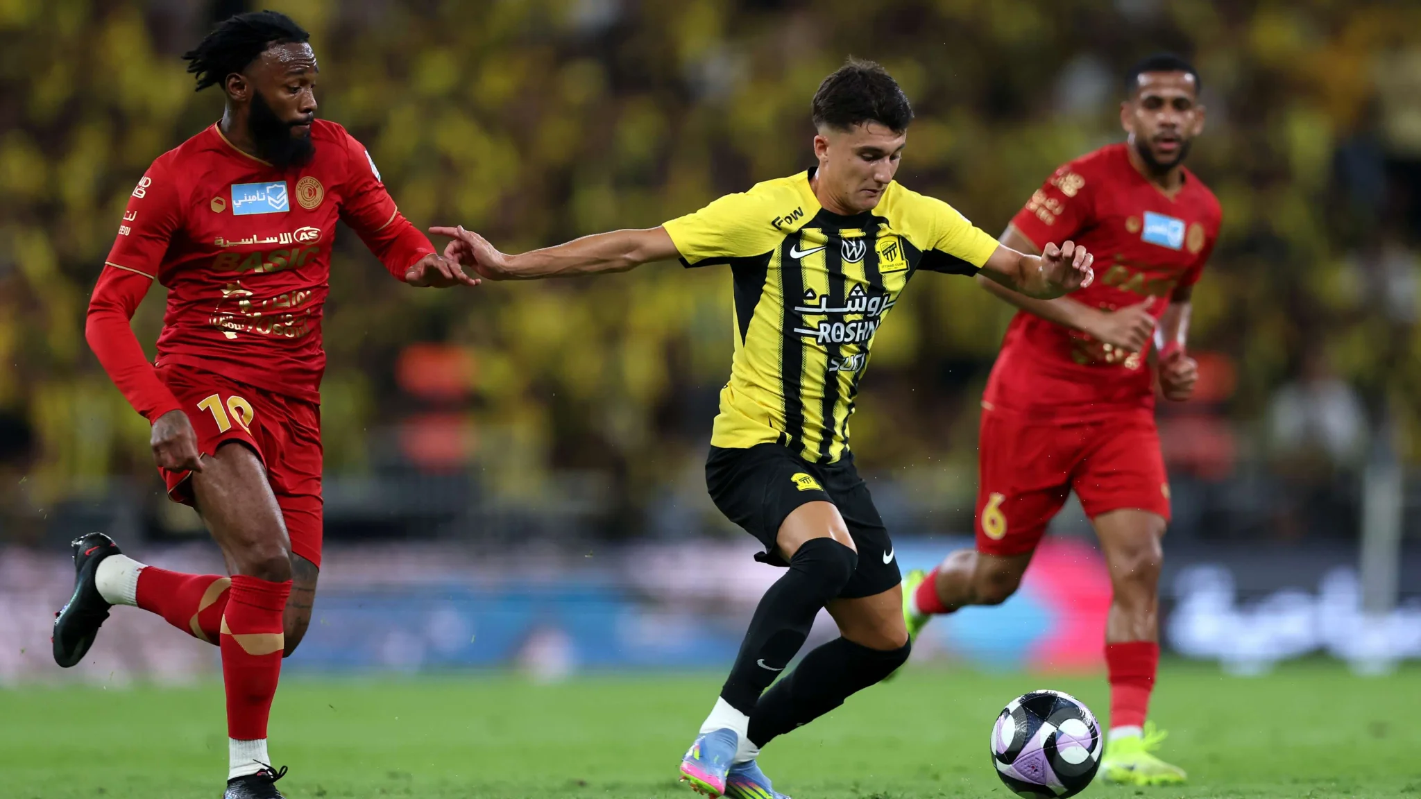 لاعب كرة قدم يرتدي زيًا أصفر من فريق Al-Shabab يتحكم بالكرة وسط لاعبين من فريق الخصم بالزي الأحمر خلال مباراة في دوري روشن السعودي.