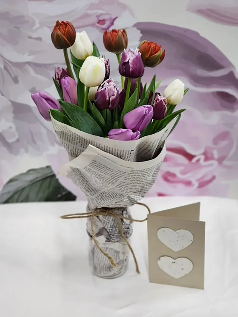 باقة من زهور التوليب الملونة موضوعة في وعاء زجاجي، محاطة بورق جرائد، مع بطاقة تهنئة مصنوعة من ورق معاد تدويره.