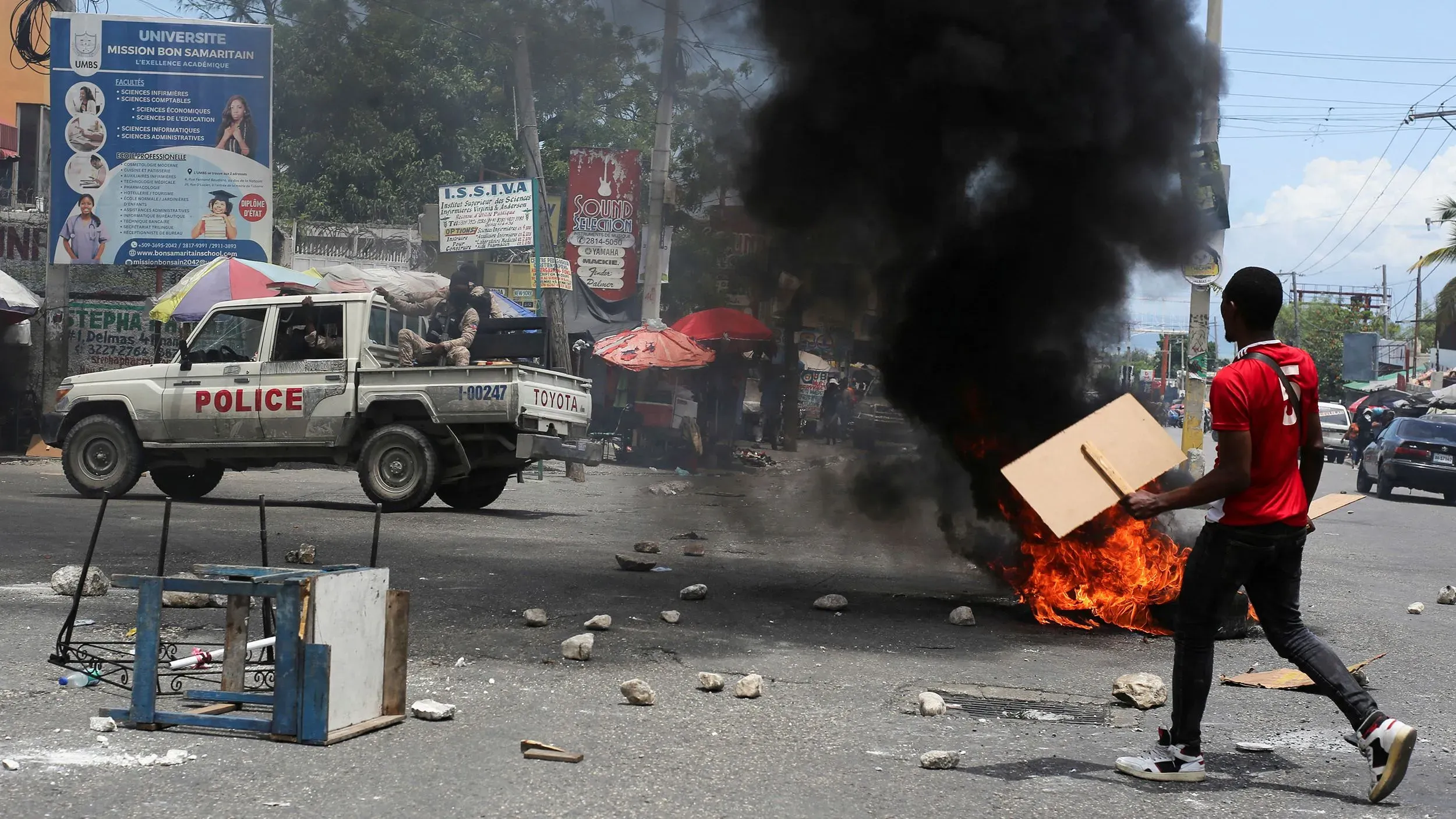 مشهد من الشارع في هايتي يظهر شابًا يحمل لافتة بينما تشتعل النيران في حطام، مع وجود سيارة شرطة في الخلفية، مما يعكس حالة الفوضى والعنف.