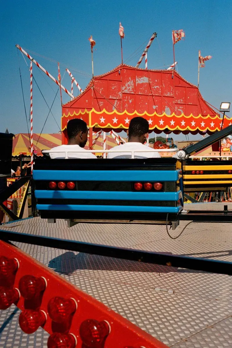 شخصان يجلسان على أرجوحة في مهرجان، مع خيمة حمراء وزخارف ملونة في الخلفية، تعكس أجواء الترفيه على الشاطئ البريطاني.