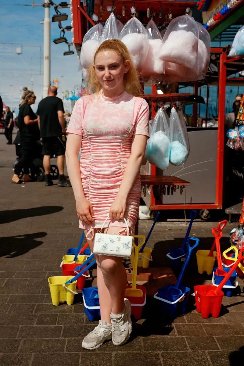 فتاة شابة ترتدي فستانًا ورديًا تقف أمام كشك يبيع حلوى القطن، مع أكياس حلوى معلقة في الخلفية وألعاب ملونة.