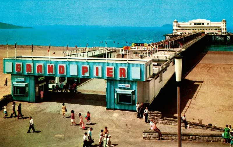 واجهة رصيف "جراند" في بريطانيا، تظهر الزوار يتجولون على الرصيف مع البحر في الخلفية، تعكس تاريخ الشواطئ البريطانية وثقافتها.