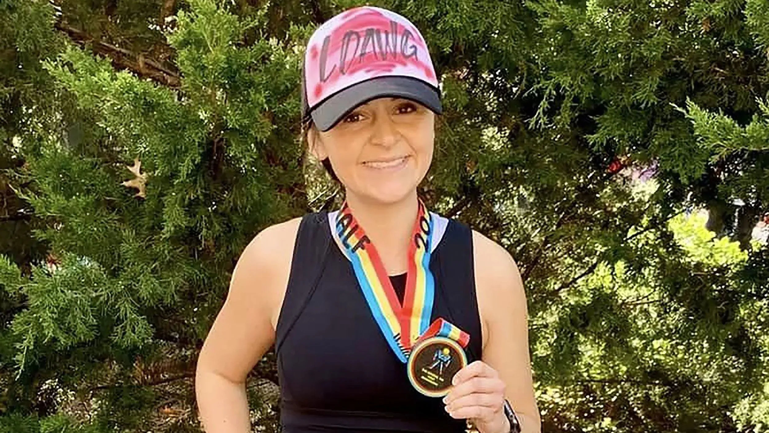 صورة لامرأة مبتسمة ترتدي قبعة تحمل كلمة \"LDANG\" وتستعرض ميدالية ملونة، تعكس إنجازاتها في سباقات الجري.