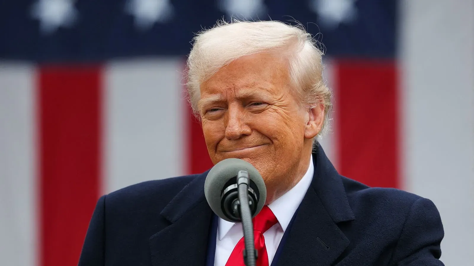 ترامب يتحدث في مؤتمر صحفي، مبتسمًا، مع العلم الأمريكي خلفه، مع التركيز على الرسوم الجمركية وتأثيرها على الاقتصاد.