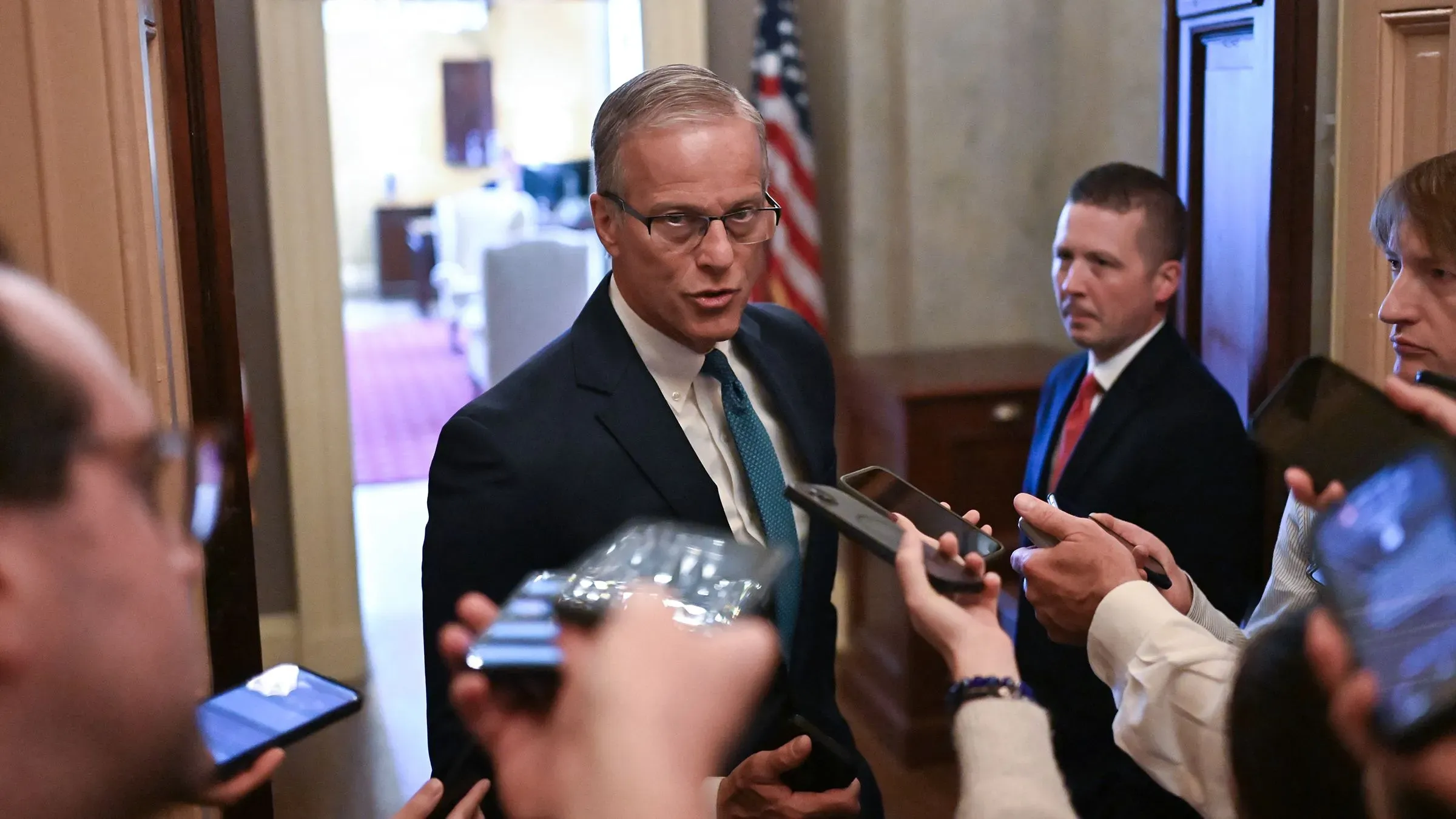 سيناتور يتحدث إلى الصحفيين في ممر مجلس الشيوخ، محاطًا بأفراد من وسائل الإعلام، وسط أجواء من التوتر حول مشروع قانون ترامب.