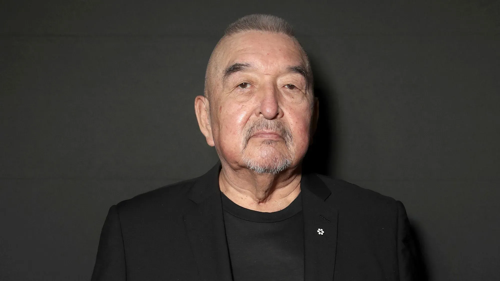 تظهر الصورة غراهام غرين، ممثل من السكان الأصليين، يرتدي بدلة سوداء، مع تعبير جاد، في خلفية داكنة. اشتهر بأدواره السينمائية والتلفزيونية.