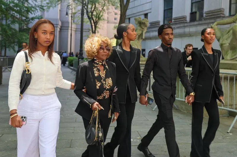 مجموعة من الأشخاص يرتدون ملابس سوداء، يسيرون معًا في منطقة حضرية، مع وجود امرأة مسنّة في الوسط.