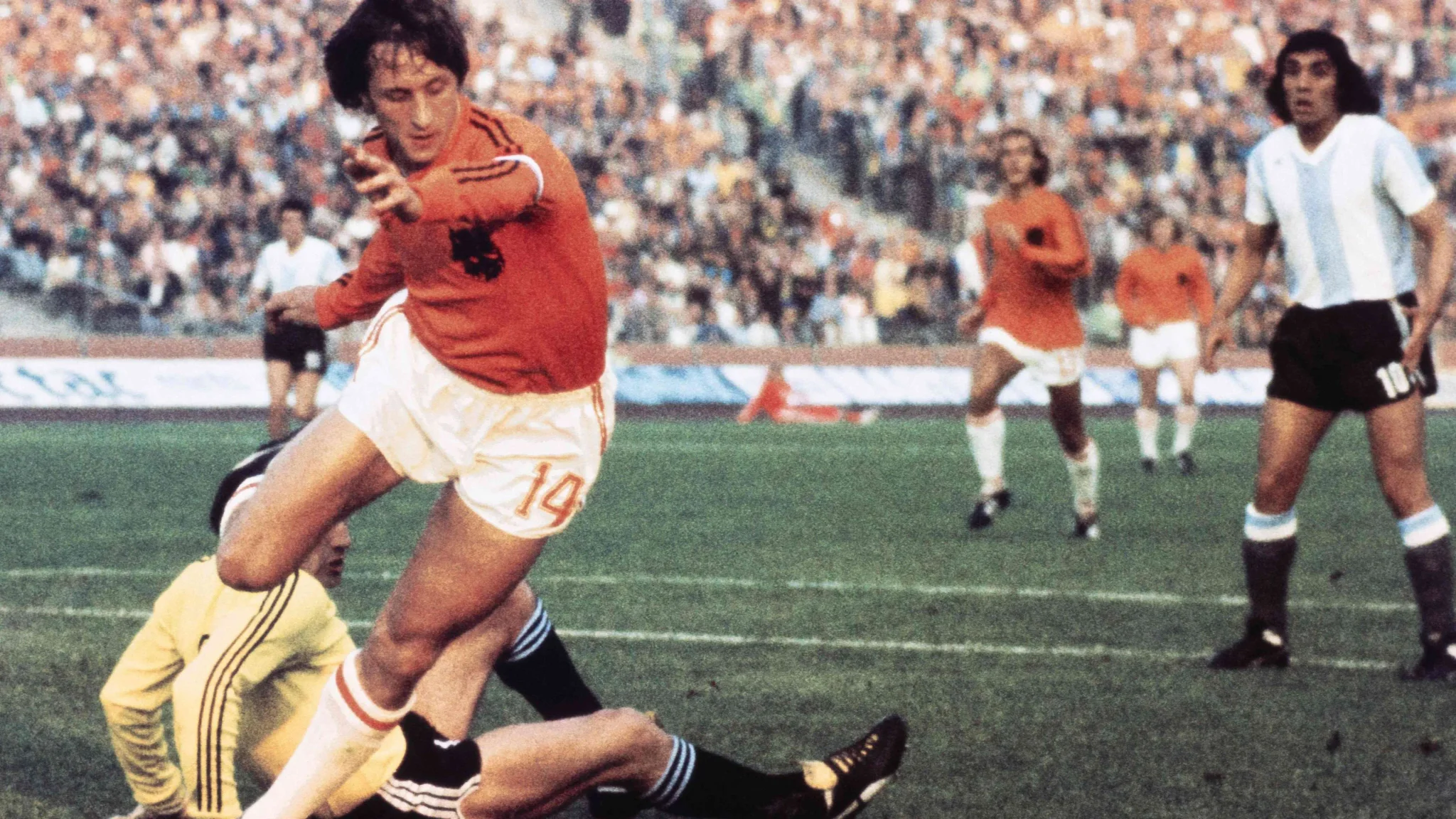 لاعب هولندي يرتدي قميصًا برتقاليًا يراوغ حارس مرمى أرجنتيني في مباراة كأس العالم 1974، مع لاعبين آخرين في الخلفية.