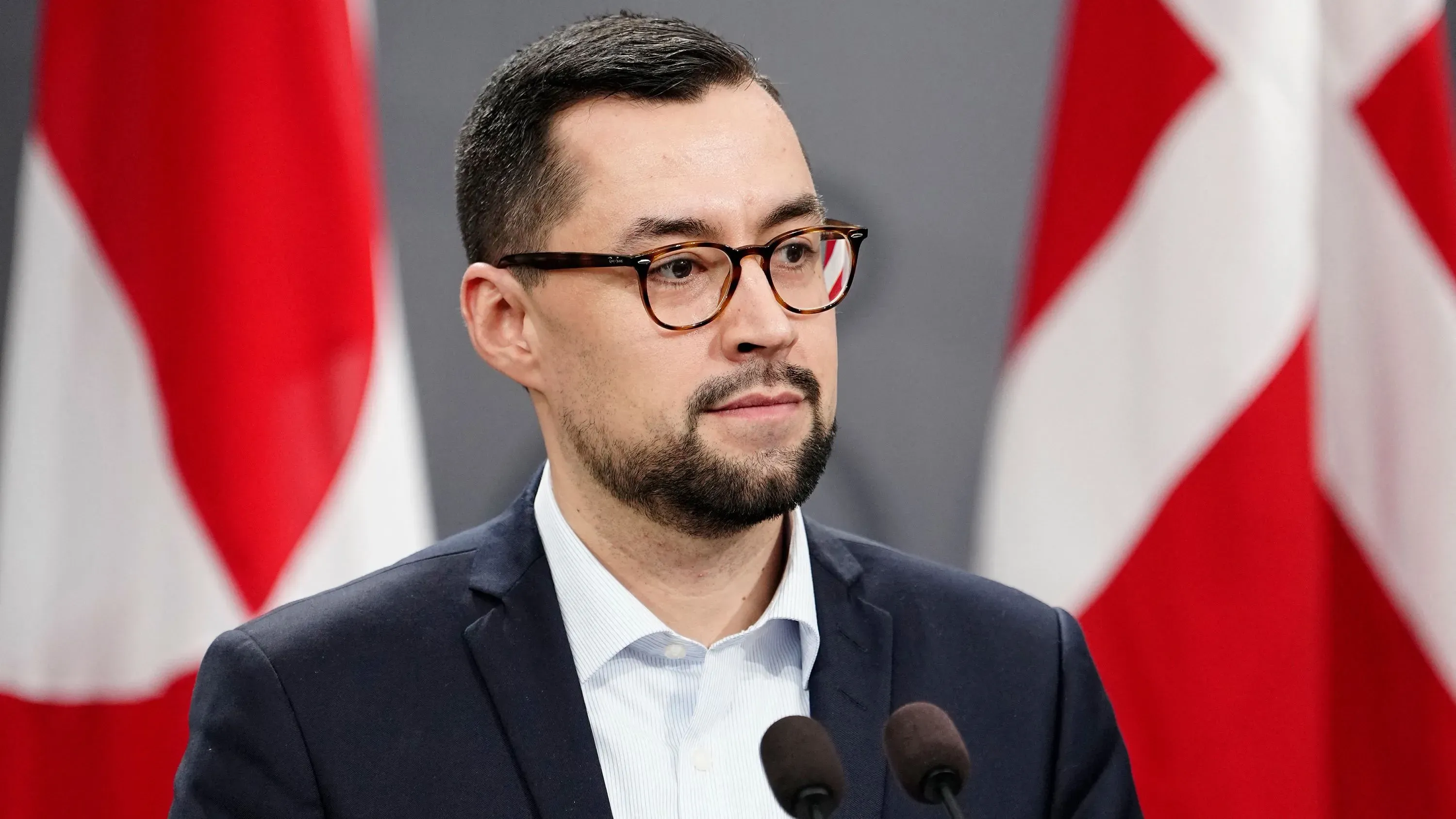 زعيم غرينلاند يتحدث في مؤتمر صحفي، مؤكدًا رغبة بلاده في الاستقلال واحترام حقوقها، مع وجود علمين خلفه.