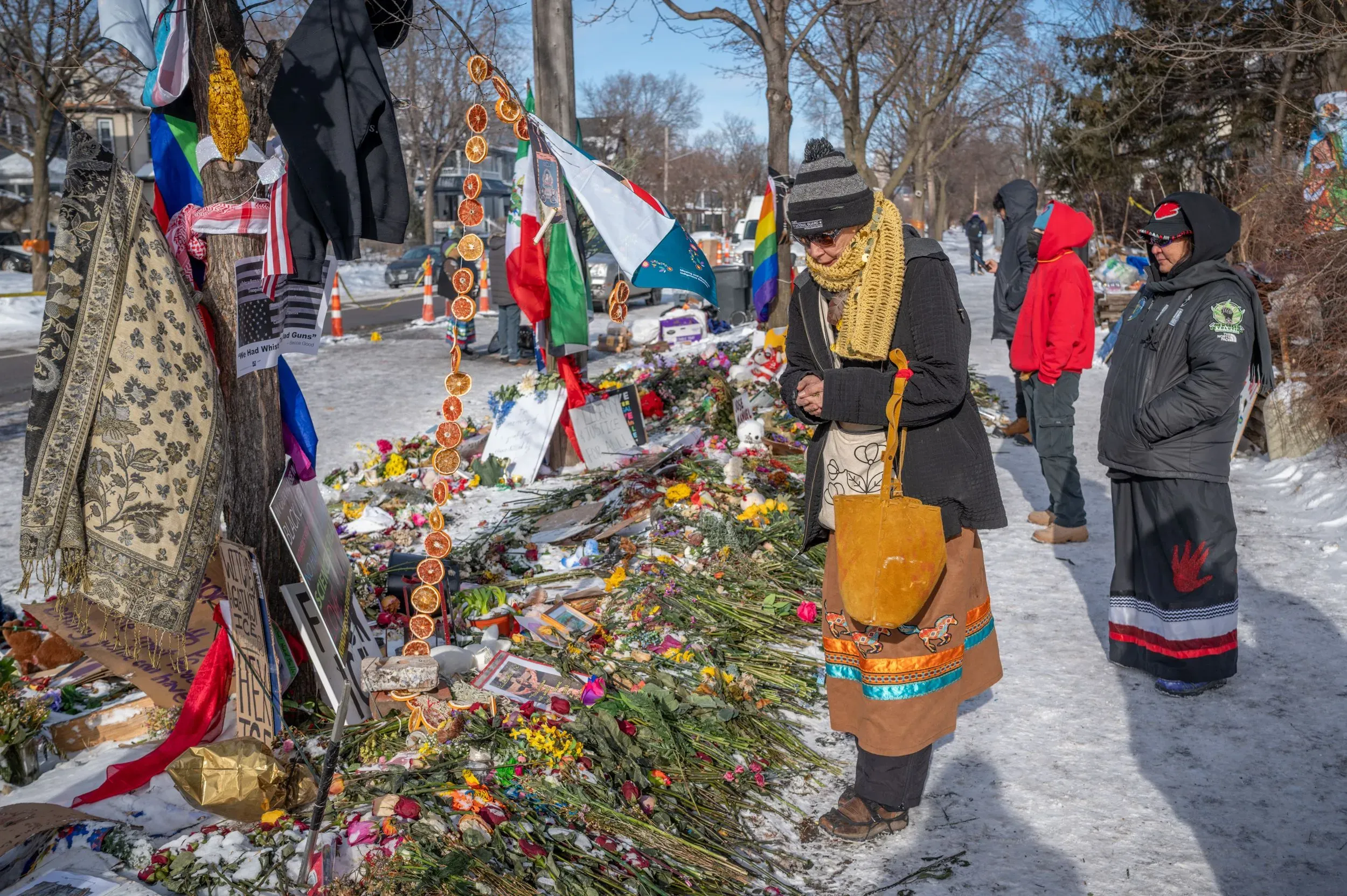موقع تذكاري يضم زهورًا وصورًا، مع أشخاص يعبّرون عن حزنهم. يظهر العلم الأمريكي وأعلام أخرى، في مشهد يعكس التأمل والذكرى.