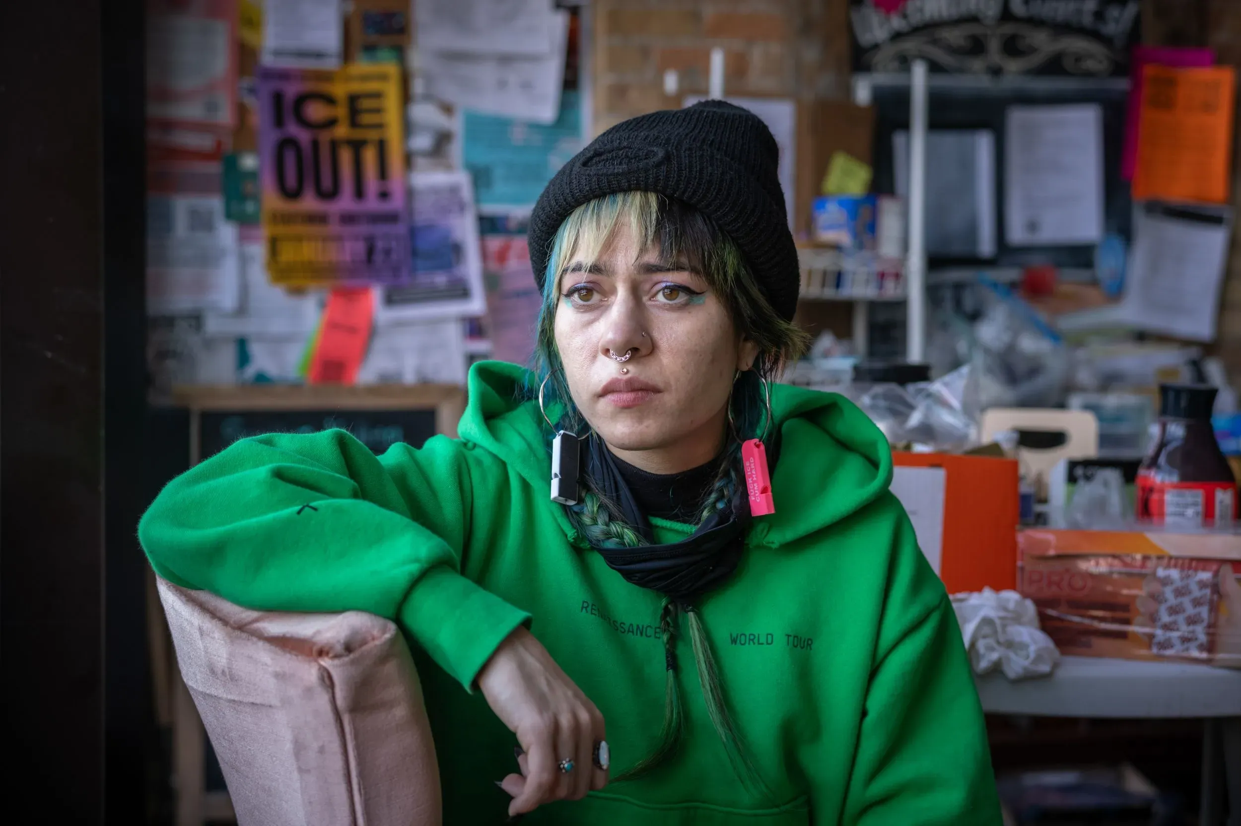 امرأة شابة ترتدي سترة خضراء وقبعة سوداء، تجلس في مكان داخلي، تعكس ملامح وجهها مشاعر التأمل والقلق، مع خلفية مليئة بالملصقات.