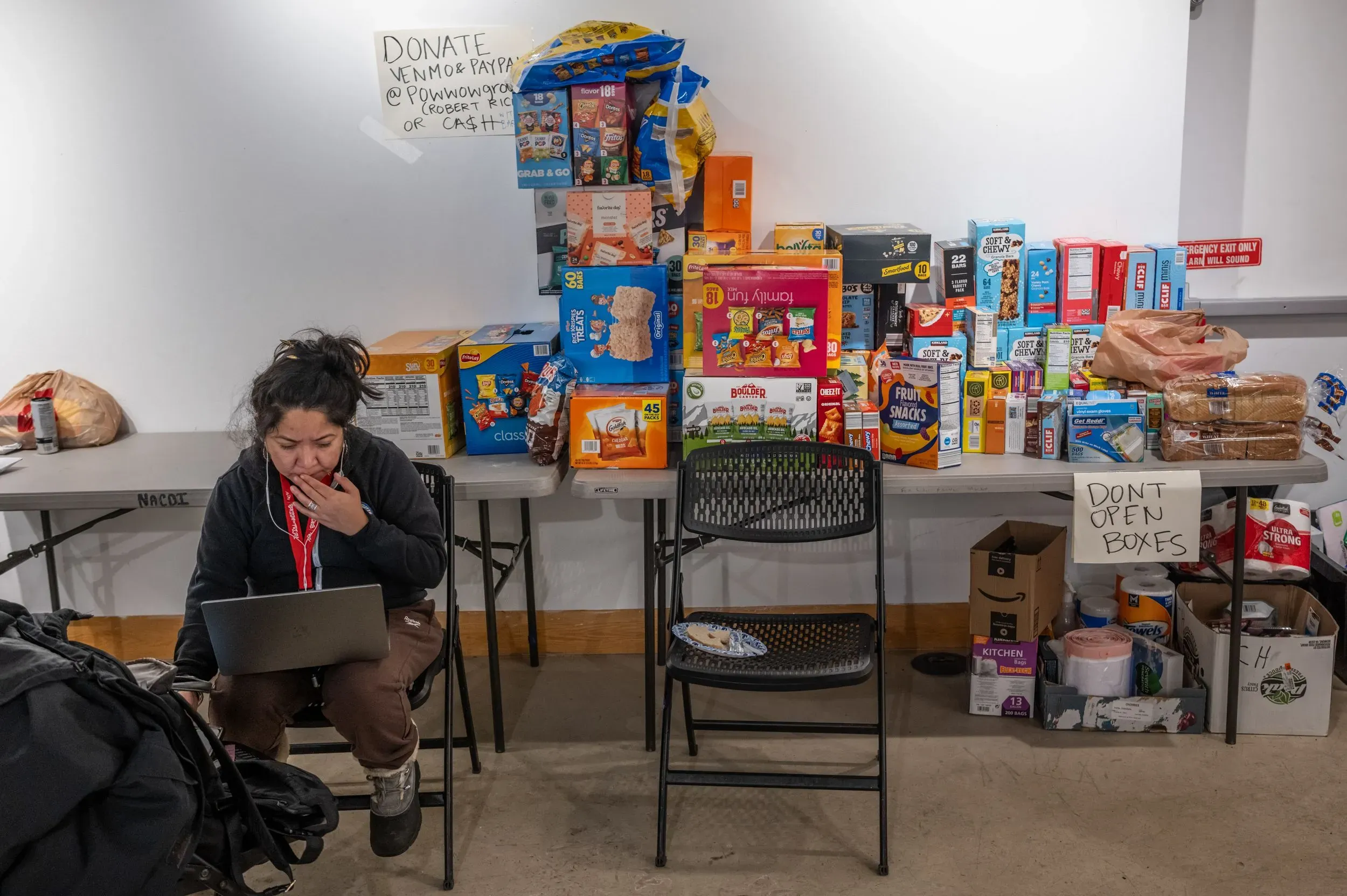امرأة تجلس أمام طاولة مليئة بالمواد الغذائية، تستخدم الكمبيوتر المحمول، بينما تظهر لافتات تطلب التبرعات.