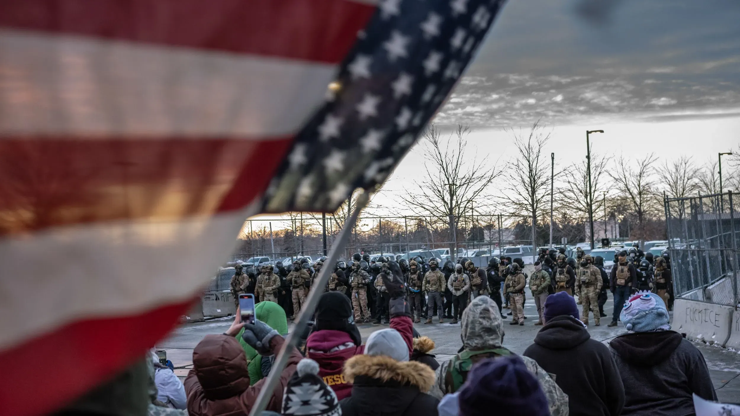 تجمع حشود من الناس أمام قوات الأمن في مينيابوليس، مع العلم الأمريكي يرفرف في المقدمة، في أجواء مشحونة بالتوتر بعد الأحداث الأخيرة.