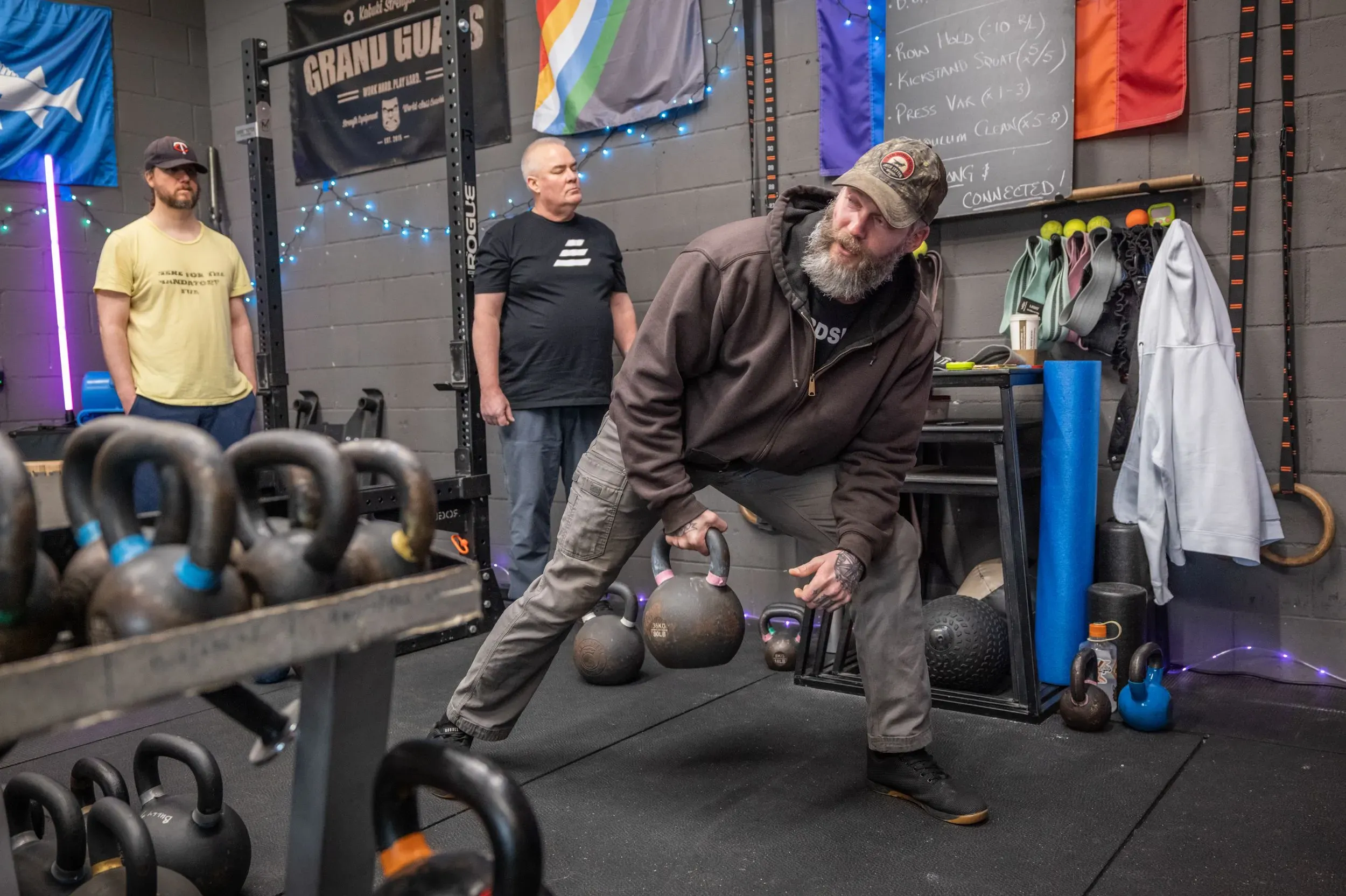 رجل ذو لحية طويلة يرتدي سترة داكنة يقوم بتمرين رفع الأثقال في صالة رياضية، بينما يشاهد اثنان من المتدربين الآخرين.