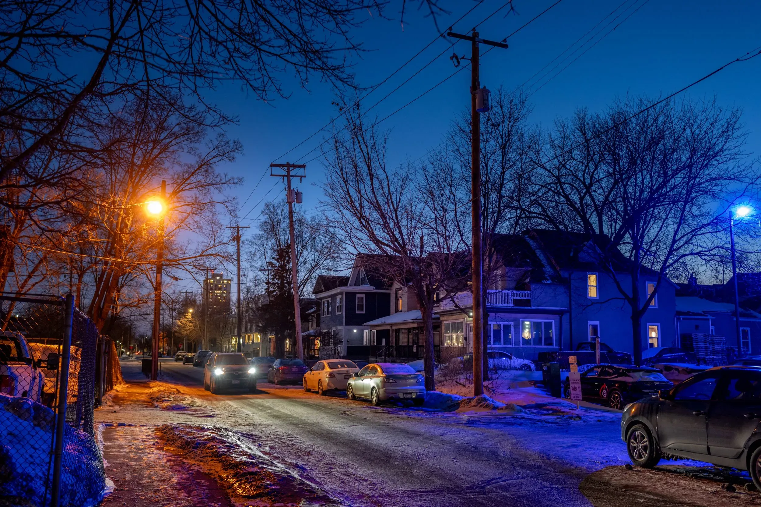 شارع في مينيابوليس مضاء بأضواء الشوارع، مع سيارات متوقفة وأشجار عارية، يعكس أجواء الشتاء القارص والتوترات الاجتماعية.