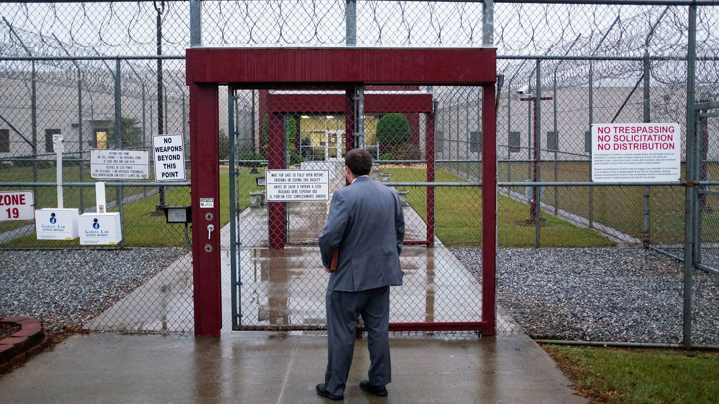 رجل يرتدي بدلة يقف أمام بوابة مغلقة لمركز احتجاز، محاط بأسلاك شائكة ولافتات تحذر من دخول الأسلحة.