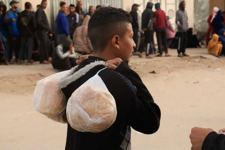 طفل يحمل خبزًا في أكياس على ظهره، منتظرًا في طابور طويل أمام مخبز في غزة، حيث يعاني السكان من نقص حاد في الدقيق بسبب القيود الإسرائيلية.