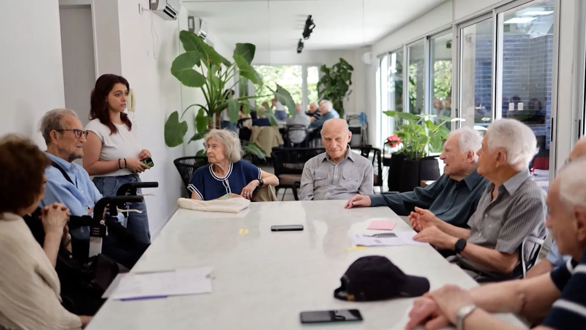 مجموعة من كبار السن يجتمعون حول طاولة في غرفة اجتماعات، مع شابة تتحدث إليهم، في إطار بودكاست "تسعون وعدّ" لمشاركة تجاربهم.
