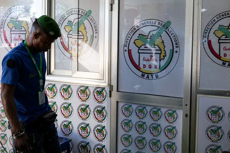 رجل يرتدي زيًا رسميًا يقف أمام مركز الانتخابات في غينيا، حيث تظهر شعارات الانتخابات على الجدران، في سياق الاستفتاء الدستوري.