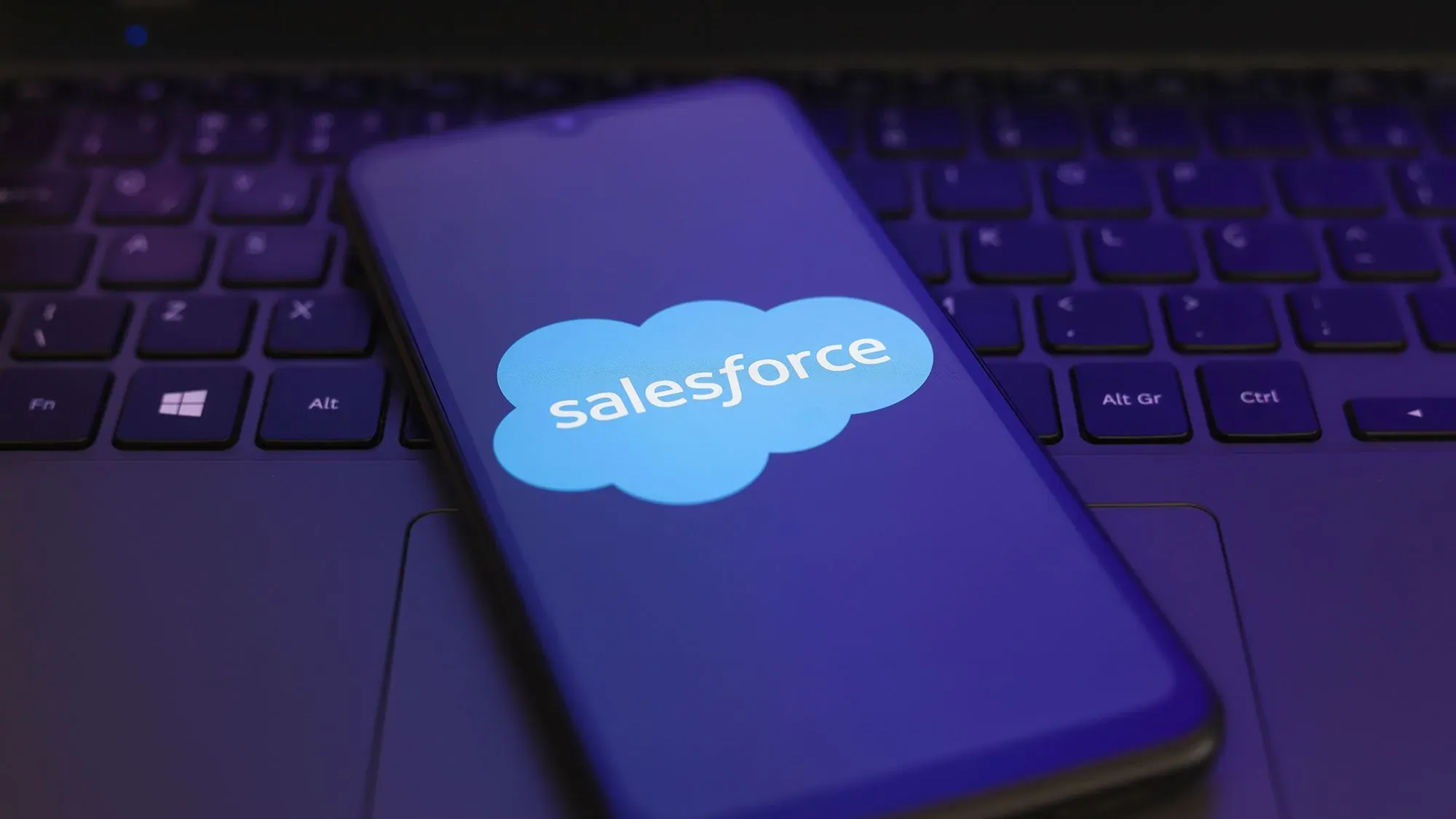 شاشة هاتف محمول تعرض شعار Salesforce بلون أزرق، موضوعة على لوحة مفاتيح كمبيوتر، تشير إلى مخاطر التطبيقات المعدلة في بيئات العمل.
