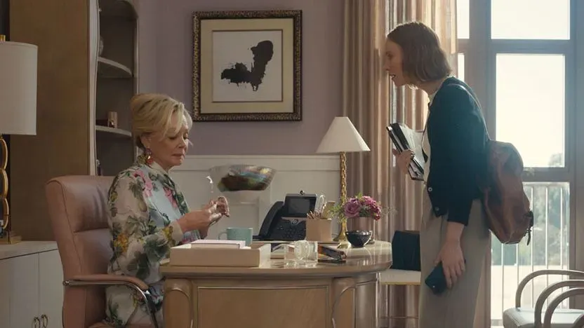 مشهد من مسلسل "Hacks" يظهر شخصيتين في مكتب؛ واحدة تجلس على مكتب أنيق بينما الأخرى واقفة تحمل ملفات، مما يعكس تفاعلهم المهني.