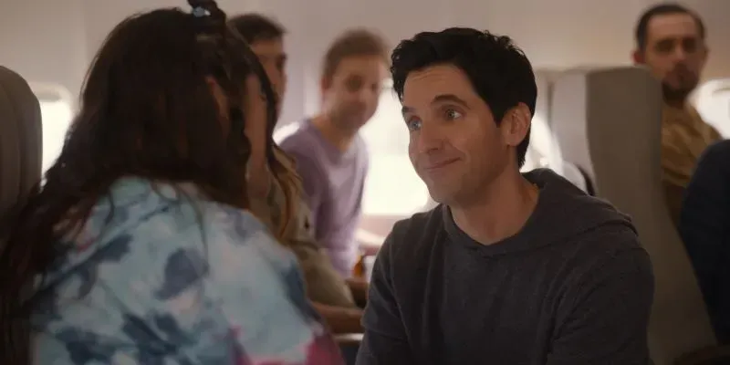 مشهد من مسلسل "Hacks" يظهر شخصية جيمي مبتسمًا أثناء حديثه مع امرأة في طائرة، مما يعكس لحظات الكوميديا والتفاعل الاجتماعي.