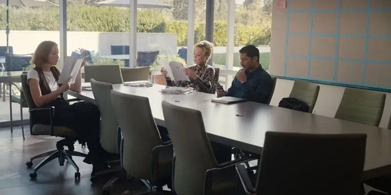 اجتماع في غرفة مؤتمرات حيث تجلس ثلاث شخصيات، اثنتان تراجعتان عن قراءة الأوراق، بينما يتفاعل الثالث مع جهاز لوحي.