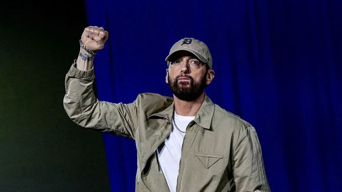 إيمينيم يرفع قبضته في عرض، مع خلفية زرقاء، معبراً عن حماسه كجد بعد إعلان ابنته هايلي عن مولودها الجديد.