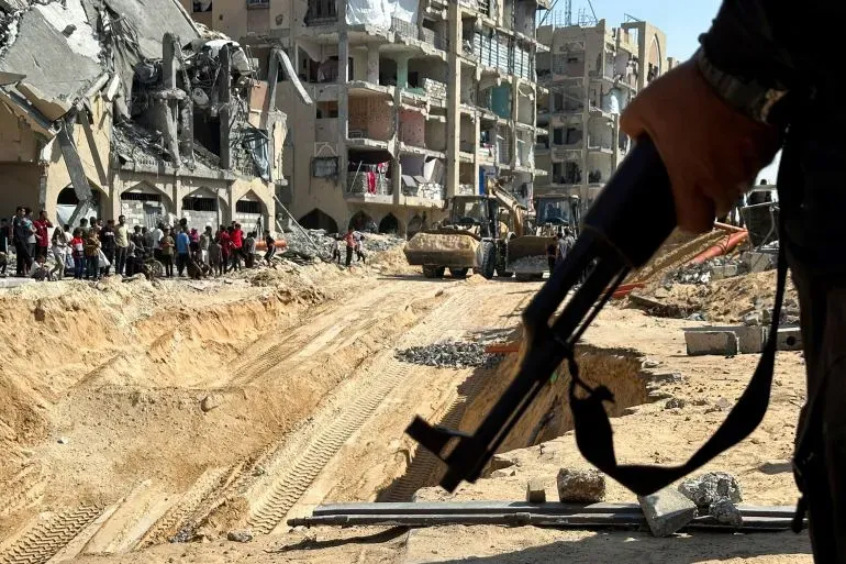 مسلح يحمل سلاحه في غزة، بينما تظهر في الخلفية المباني المدمرة وأعمال إعادة الإعمار، مع تجمع للناس في المنطقة.