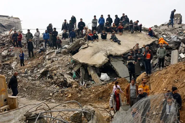 مجموعة من الأشخاص يتجمعون حول أنقاض مبنى مهدوم في غزة، يعكس تأثير الهجمات الإسرائيلية المستمرة على المدنيين.