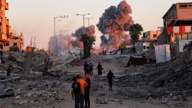 دخان يتصاعد من خلف مبانٍ مدمرة في غزة، مع وجود عدة أشخاص يتجولون في الشارع وسط الدمار، مما يعكس آثار الحرب المستمرة.