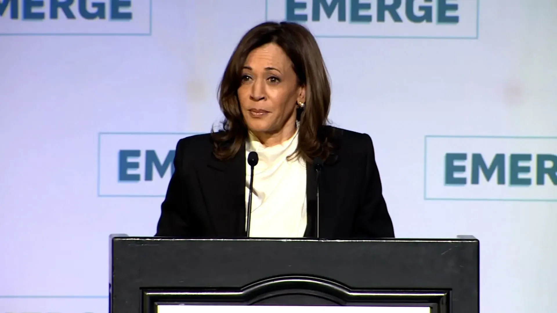 كامالا هاريس تتحدث في حفل Emerge Gala، متهمة ترامب بتأجيج أزمة اقتصادية، وداعمة للنساء في السياسة، مع وجود شعار "EMERGE" خلفها.