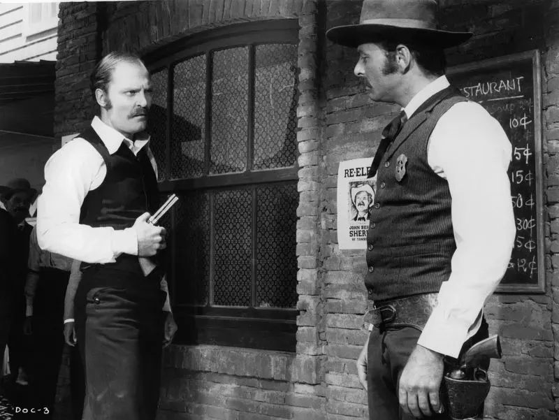 مشهد درامي يظهر رجلين يرتديان ملابس غربية تقليدية، أحدهما يحمل مسدسًا، في إطار تاريخي يعكس أجواء الأفلام الكلاسيكية.