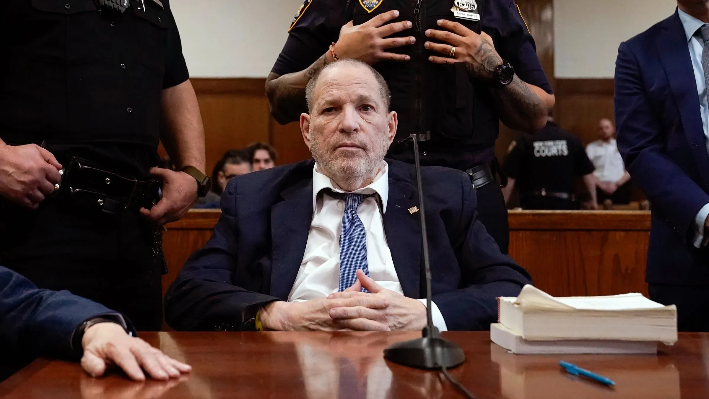 هارفي واينستين يجلس على طاولة الدفاع في المحكمة، محاطًا برجال الشرطة، ويناشد القاضي لتقديم موعد محاكمته بسبب ظروف احتجازه الصعبة.