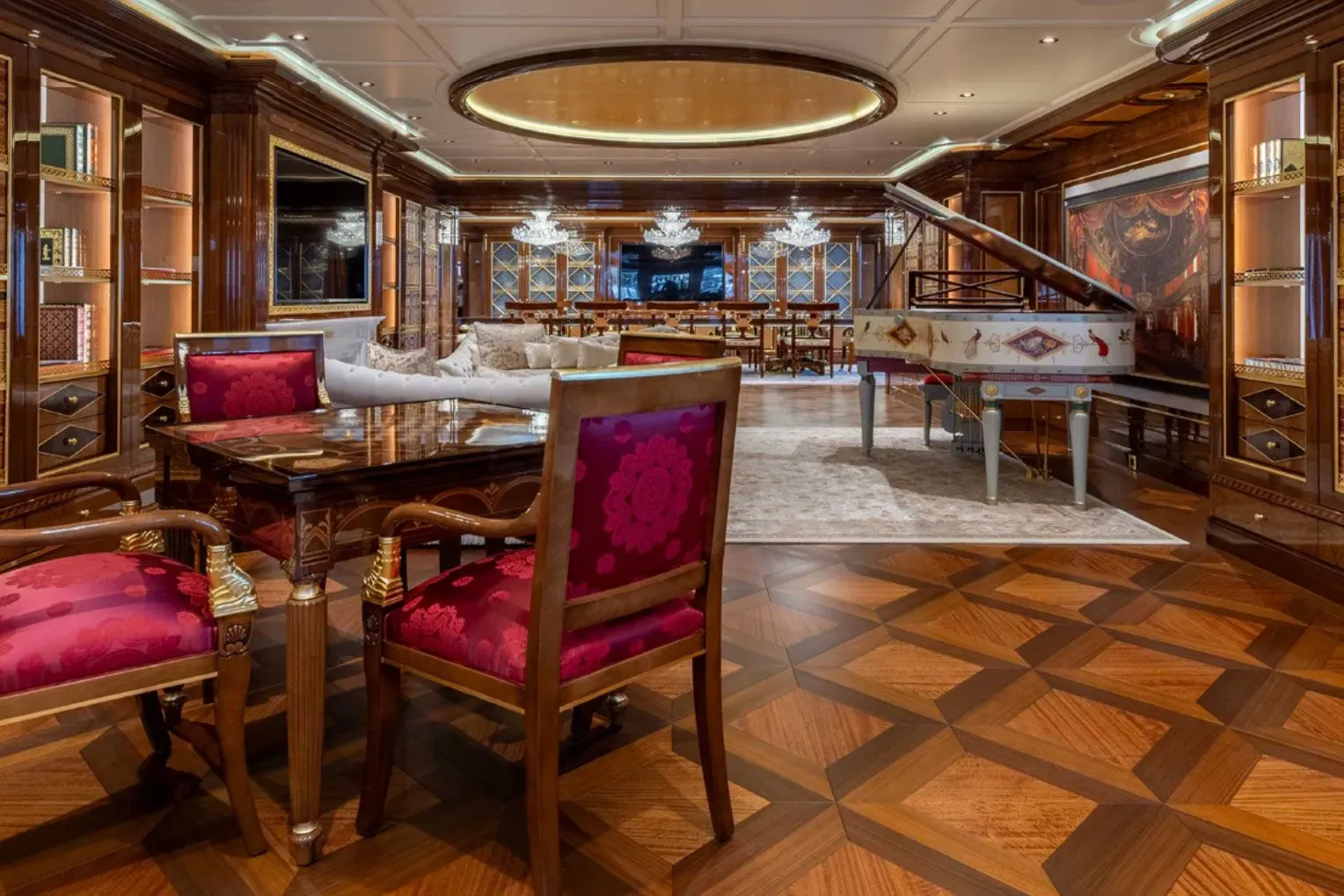 ديكور فاخر داخل يخت "أماديا" يشمل طاولة خشبية أنيقة وكراسي حمراء، مع بيانو أبيض في الخلفية، يعكس الفخامة التي يتميز بها اليخت.