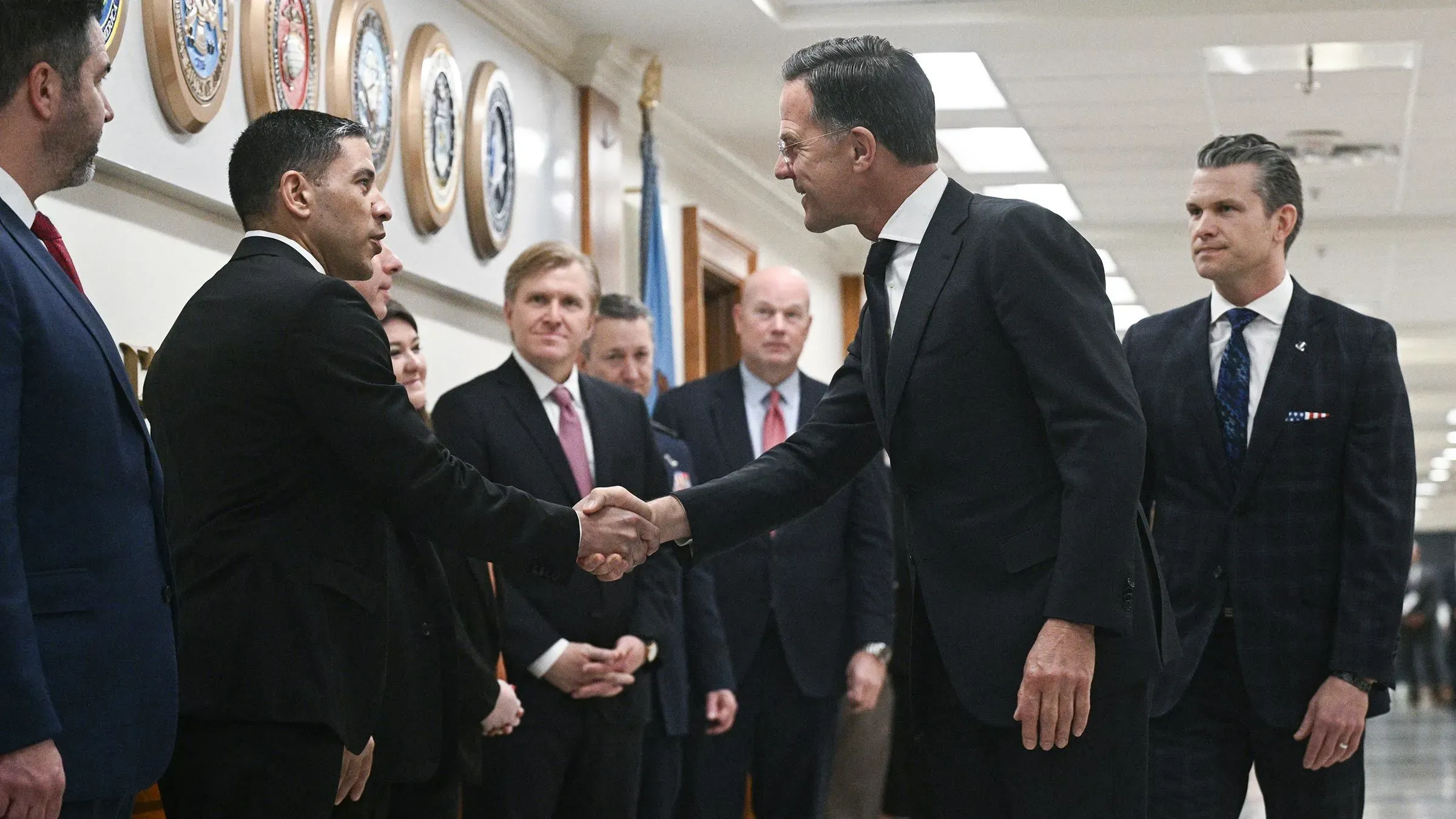 لقاء بين مسؤولين عسكريين ومدنيين في البنتاجون، حيث يتم تبادل التحيات في أجواء رسمية، مع ظهور شعارات عسكرية على الجدران.