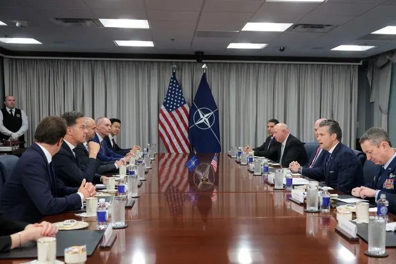 اجتماع في البنتاغون بحضور مسؤولين عسكريين ومدنيين، مع العلم الأمريكي وعلم الناتو في الخلفية، يناقش قضايا عسكرية حساسة.