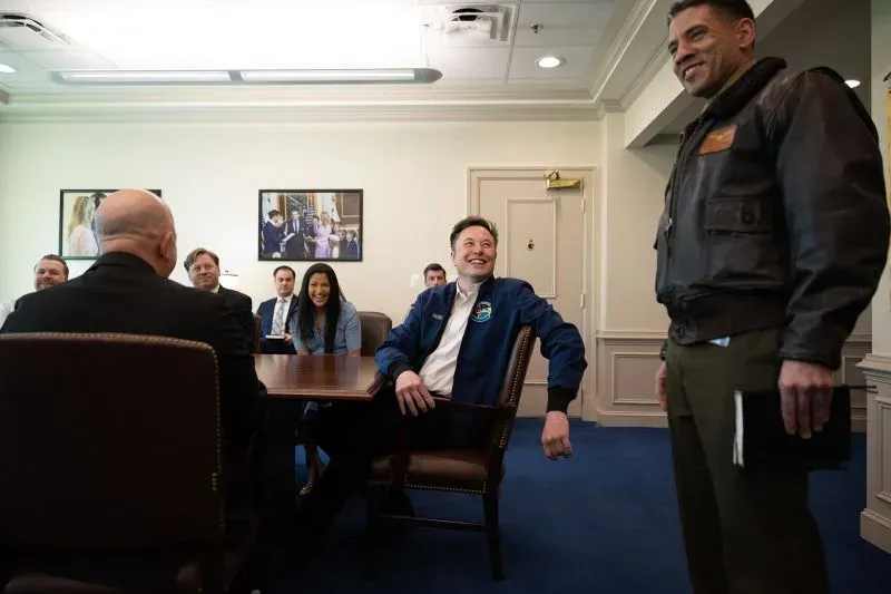 اجتماع في البنتاغون حيث يبتسم إيلون ماسك بينما يتحدث ضابط عسكري، مع وجود عدد من الحضور الآخرين في الغرفة.