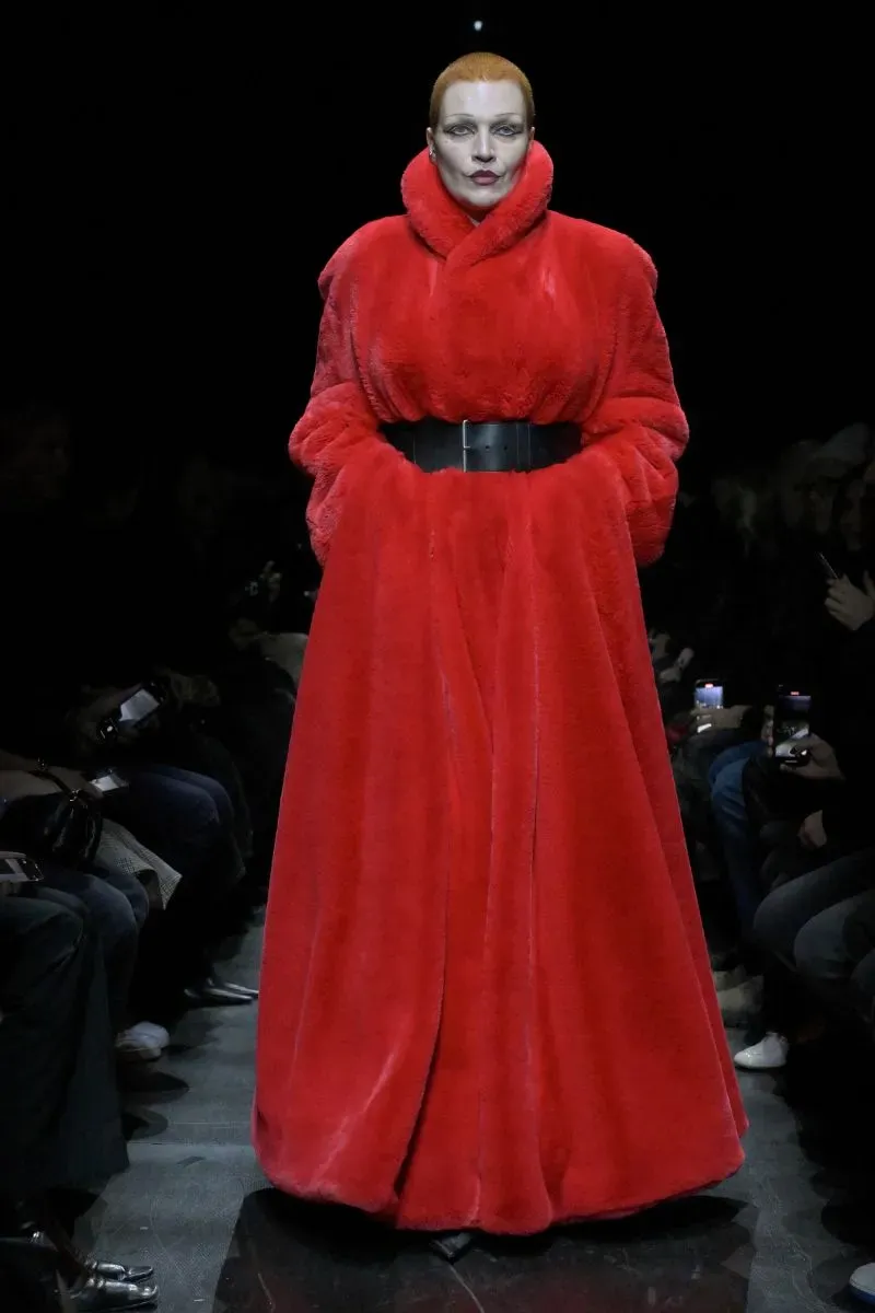 عارضة أزياء ترتدي معطفاً فخماً باللون الأحمر، مع حزام أسود، أثناء عرض أزياء في بيئة مظلمة، مما يبرز تصميم غوتشي الجديد.