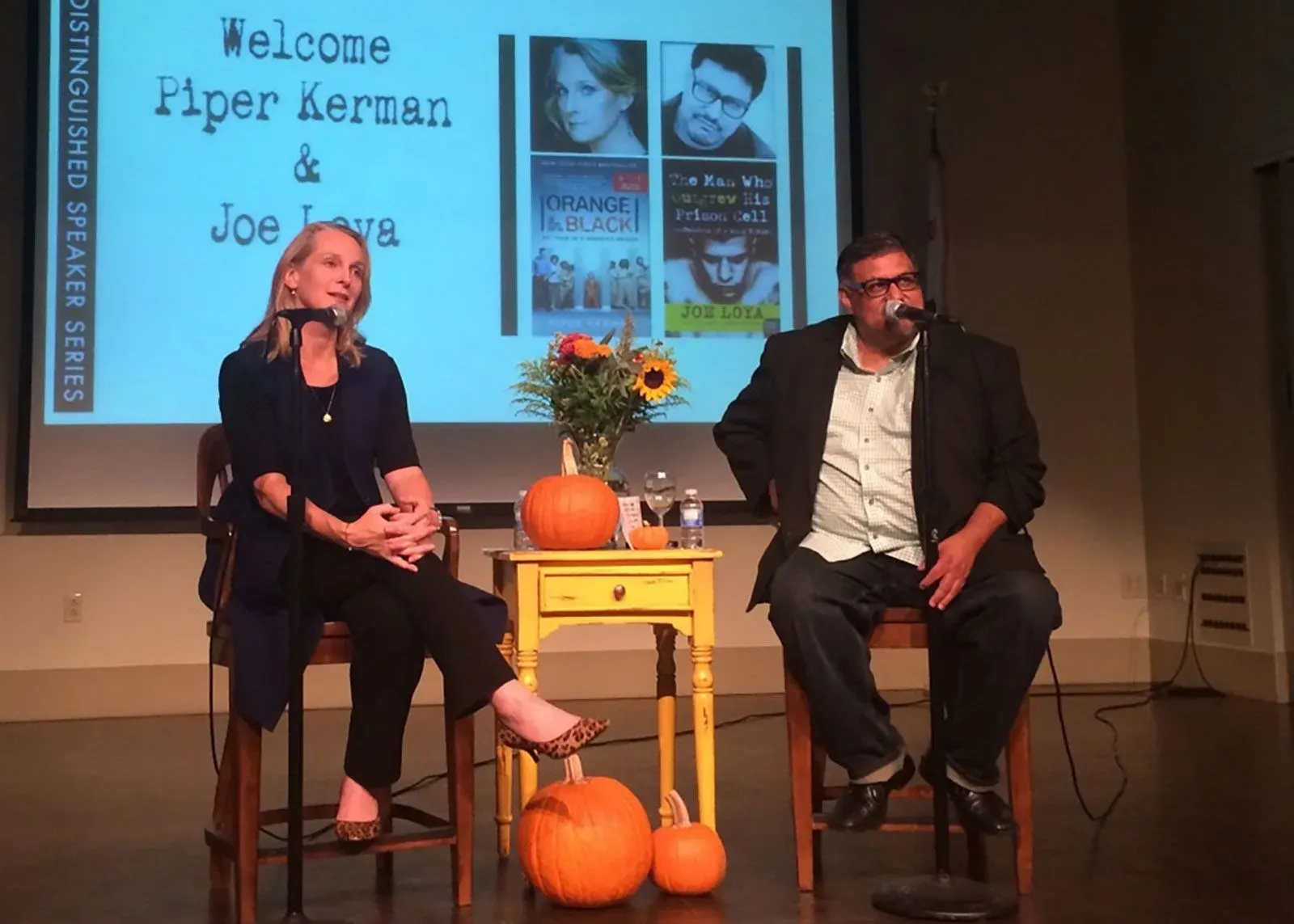 جو لويا وبايفر كيرمان يتحدثان في حدث أدبي، مع عرض كتب خلفهم، وزهور شمسية على الطاولة، وتفاح برتقالي، مما يعكس موضوع السيرة الذاتية والتغيير.