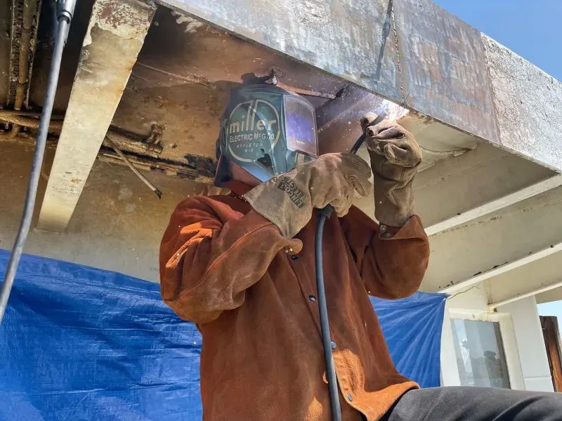 شخص يرتدي خوذة لحام ويعمل على إصلاح هيكل سفينة قديمة، تعكس جهود الترميم والتجديد للسفينة السياحية "أورورا".