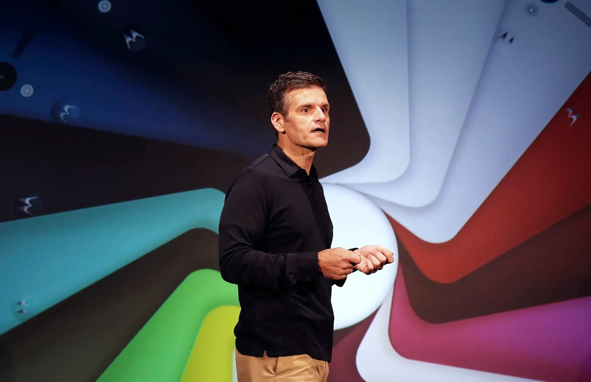 دينيس وودسايد، الرئيس التنفيذي السابق لموتورولا، يتحدث في حدث تكنولوجي، مع خلفية ملونة تعكس تصميمات هواتف ذكية.