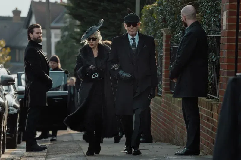 امرأة ترتدي ملابس سوداء أنيقة مع قبعة كبيرة، تسير مع رجل في بدلة سوداء، بينما يراقبهم رجال آخرون في ملابس رسمية، في مشهد من مسلسل "موب لاند".